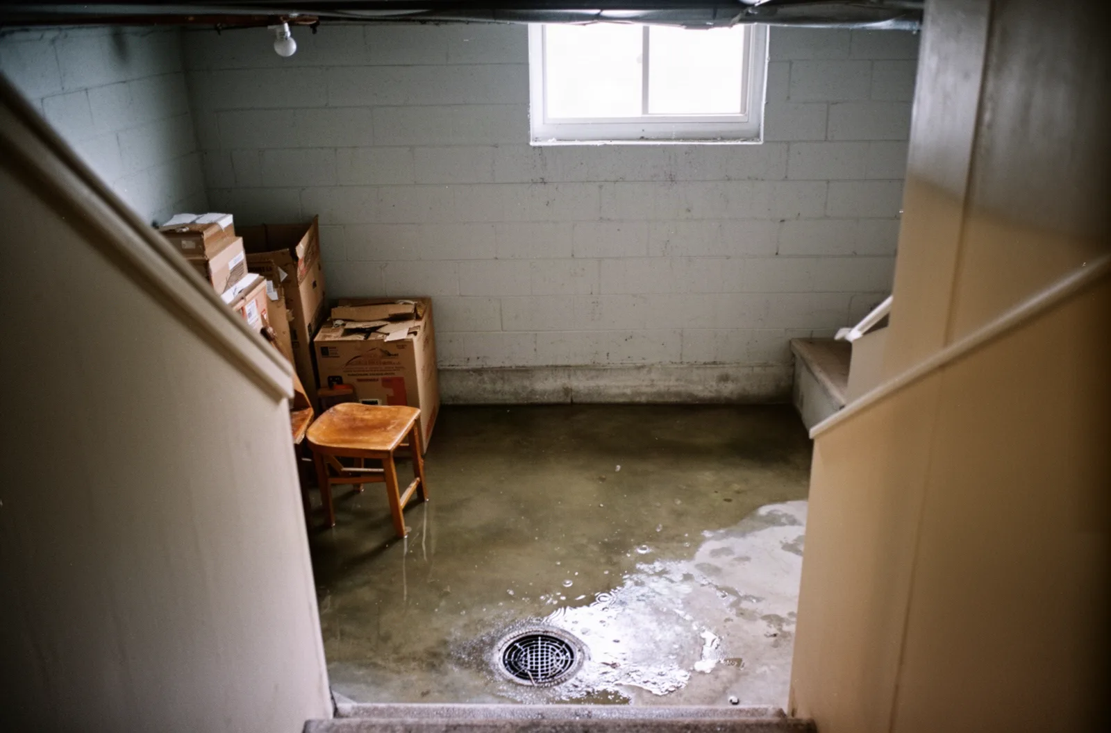 Standing water on a residential basement floor with boxes and a floor drain, seen from the stairs